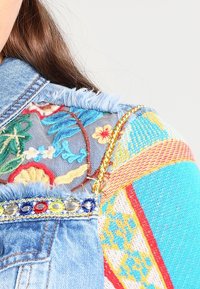 Denim takki, jossa on värikkäitä brodeerattuja kukkakuvioita ja kudottu monivärinen kangaspaikka, yllä henkilö, jolla on ruskeat hiukset.