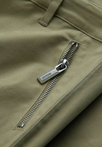 Gros plan sur un tissu vert olive avec une fermeture éclair en métal et une languette étiquetée "onore" sur une poche de vêtement.