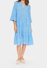 Ljusblå knälång klänning med vita prickar, V-ringning och trekvartslånga klockärmar. Har en lager-på-lager kjol och lättviktig tyg.