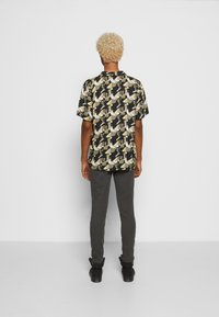 Camicia a maniche corte nera con un motivo geometrico crema e giallo, abbinata a pantaloni grigi e scarpe nere. Tessuto strutturato, vestibilità comoda.