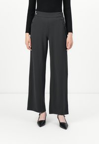 Donna che indossa pantaloni larghi neri con dettagli a pulsante in vita e décolleté neri a punta, in piedi contro uno sfondo bianco semplice.