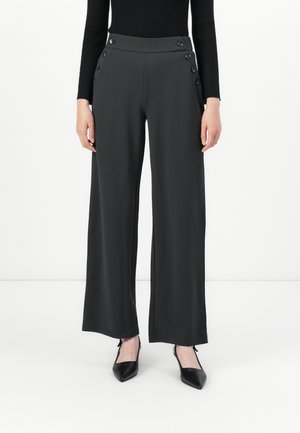 Donna che indossa pantaloni larghi neri con dettagli a pulsante in vita e décolleté neri a punta, in piedi contro uno sfondo bianco semplice.
