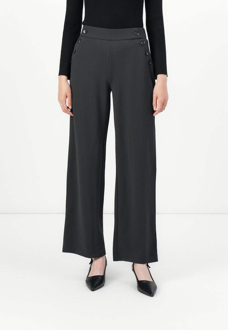 Donna che indossa pantaloni larghi neri con dettagli a pulsante in vita e décolleté neri a punta, in piedi contro uno sfondo bianco semplice.