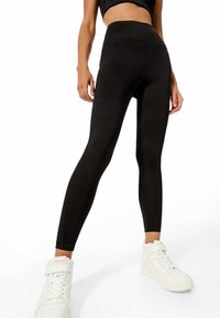 Leggings noirs taille haute avec une texture lisse, présentant un design ajusté et une construction sans coutures. Associés à des baskets montantes blanches.