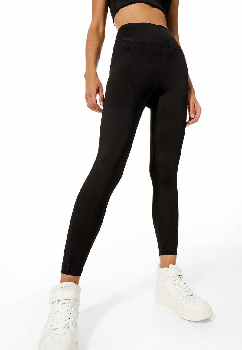Leggings noirs taille haute avec une texture lisse, présentant un design ajusté et une construction sans coutures. Associés à des baskets montantes blanches.