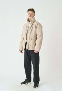 Beige gepolsterte Jacke mit hohem Kragen, Frontreißverschluss und zwei großen Taschen; getragen über einem weißen Hemd und kombiniert mit schwarzen Hosen.