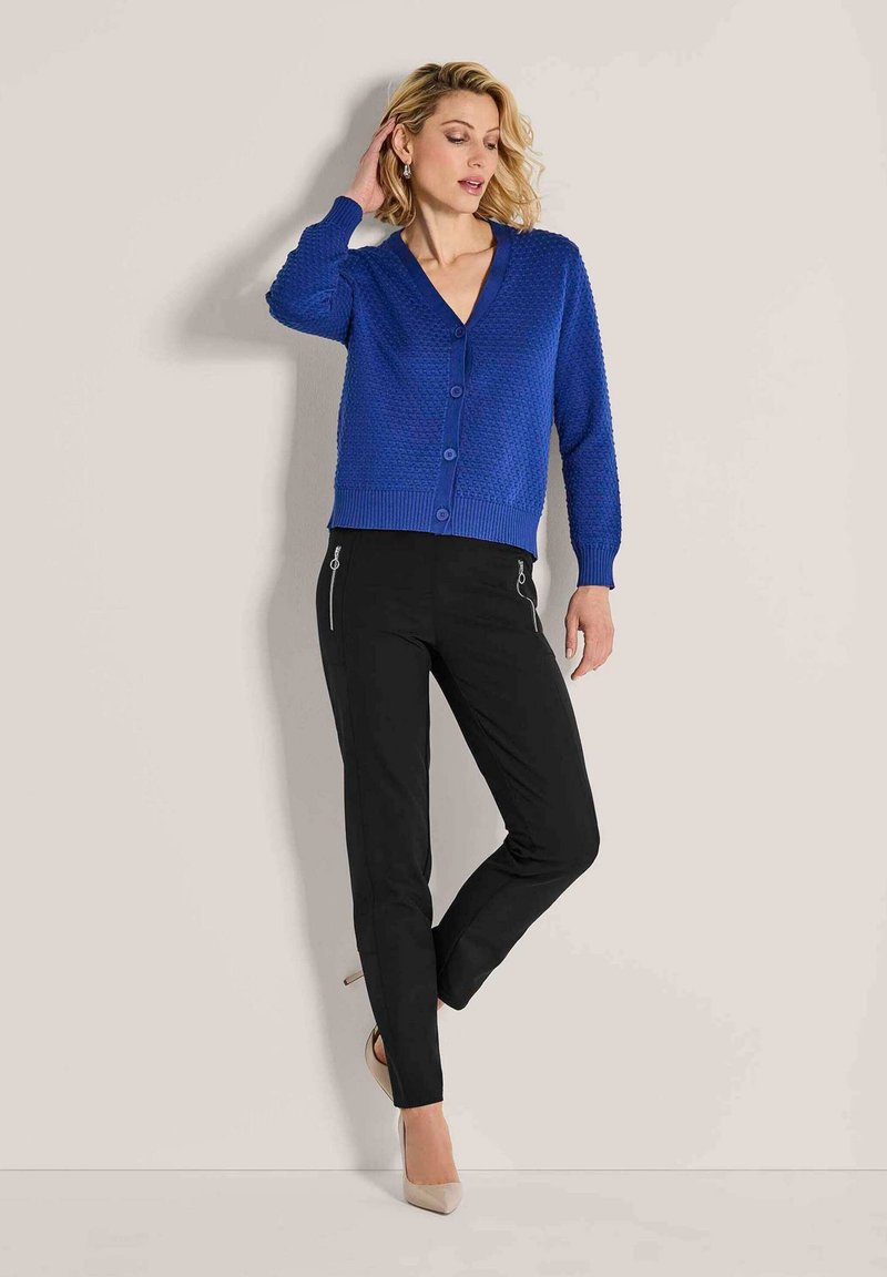 Blauwe gebreide cardigan met een V-hals en knoopsluiting, gecombineerd met zwarte broek met ritsen, gedragen met nude hoge hakken.