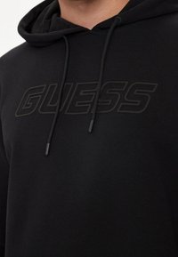 Hoodie nera con un grande logo 'GUESS' testurizzato in nero sul petto. Presenta un cappuccio con cordino e polsini a coste. Tessuto morbido.