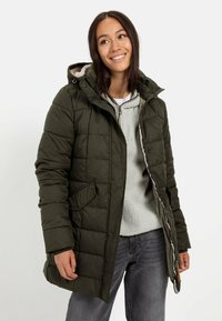 Oliven grøn puffjakke med quiltet design, h�ætte og sidelommer, iført over en lys grå fleece. Jakken har en frontlynlås og en afslappet pasform.