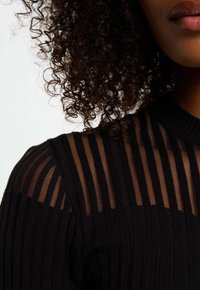 Top in maglia nera con pannelli a strisce trasparenti sulle spalle. Presenta una trama a coste e un collo rotondo, evidenziando il contrasto tra i tessuti.