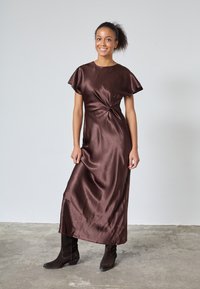 Kvinna som står inomhus iklädd en glänsande brun satin-klänning med en sidoknopt och svarta mockaankelstövlar på ett betonggolv.