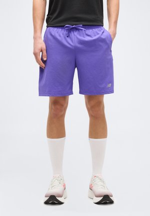 Άνδρας που φοράει μωβ σορτς New Balance, λευκές κάλτσες και ανοιχτόχρωμα παπούτσια τρεξίματος, στέκεται μπροστά σε απλό φόντο.