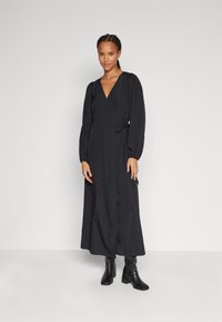 Rochie neagră, cu mâneci lungi, de tip wrap, cu decolteu în V și cordon la talie. Material neted, cu tiv până la gleznă, asortată cu cizme negre cu toc.