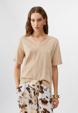 Giovane donna con capelli mossi che indossa una maglia beige a scollo a V con strass e pantaloni larghi a gamba larga con motivi nei toni del marrone e del giallo.