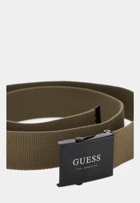 Maslinasto zeleni pojas od mrežastog materijala s crnim metalnim kopčanjem i srebrnim logotipom "GUESS". Glatka tekstura i prilagodljiva dužina.