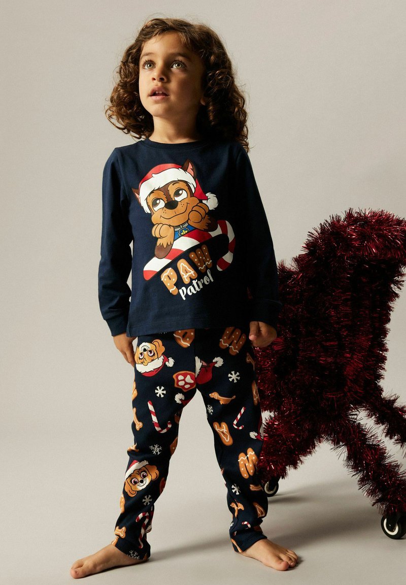 Pajama set za djecu s temom Paw Patrol: majica dugih rukava u mornarskoj plavoj boji s dizajnom crtanog psa; odgovarajuće hlače sa šarenim prazničnim uzorcima.