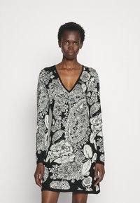 Zwarte jurk met lange mouwen, V-hals, met een zwart-wit bloemen paisley patroon. Gebreide textuur, knielang ontwerp.