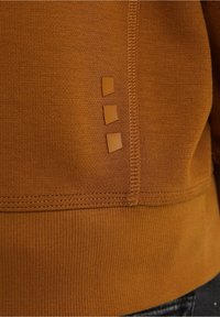 Tissu brun texturé avec trois petites patches carrées en caoutchouc disposées verticalement près d'une couture cousue et une ceinture côtelée.