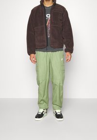 Brun fleecejacka med dragkedja, gröna cargo-byxor med fickor och svarta sneakers med vita och gröna detaljer. Grå t-shirt med grafik.