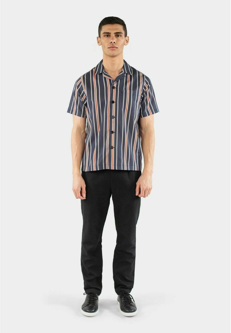 Kortærmet skjorte med navy bund og lodrette striber i orange og hvid, knappet fortil, parret med sorte bukser og sorte sko.