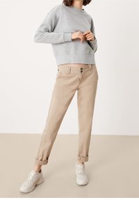 Osoba v šedé mikině, béžových ohrnutých kalhotách a bílých teniskách stojící před jednobarevným pozadím.