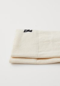 Cream-colored folded knit fabric with a small black fish symbol embroidered near the edge on a white background.