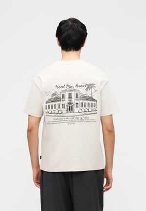 Άτομο που φοράει λευκό μπλουζάκι με μαύρη γραμμή σχεδίου ενός ξενοδοχείου και φοίνικα, και κείμενο που αναγράφει "Hotel Mar Bravo" στην πλάτη.