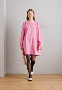 Ροζ φόρεμα με μακριά μανίκια, με χαλαρή εφαρμογή και δύο μπροστινές τσέπες, συνδυασμένο με μαύρες κολάν και λευκές μπαλαρίνες. Λείος υφασμάτινος ιστός.