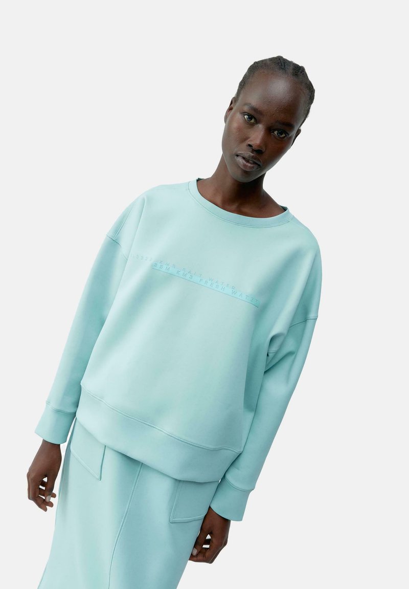 Felpa leggera color teal con una vestibilità comoda, collo a giro e polsini a costine. Presenta un design testuale in rilievo sul petto e una superficie liscia.
