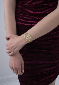 Orologio d'oro con cinturino a rete, quadrante quadrato e dettagli argentati. Indossato su una mano, con una finitura liscia su un vestito testurizzato bordeaux.