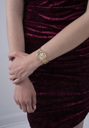 Montre en or avec un bracelet en maillage, boîtier carré et accents argentés. Portée sur une main, avec une finition lisse contre une robe texturée bordeaux.