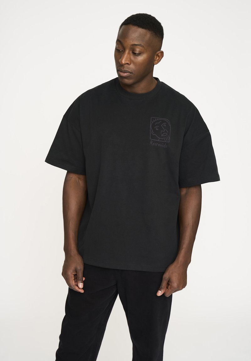 Sort kortærmet bomuldstrøje med rund halsudskæring, der har et lille grafisk logo på venstre side, der viser en ansigtskontur.