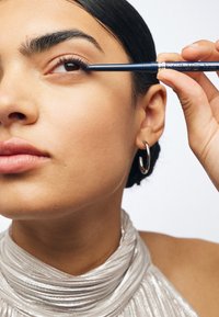 Niebieski żelowy eyeliner nałożony na powiekę przez osobę z naturalnym makijażem, ubraną w srebrny materiał o teksturze i srebrne kolczyki w kształcie kół. Gładka skóra.