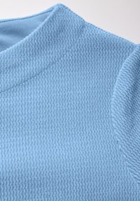 Svetle modrá texturovaná tkanina zobrazuje detailní pohled na výstřih topu. Materiál má žebrovaný vzor a jemnou, mírně chlupatou texturu.