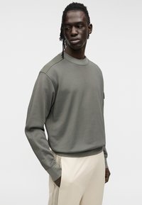 Πράσινο-ελαιί πράσινο φούτερ με στρογγυλή λαιμόκοψη και ριμπ μανσέτες, με ομαλή υφή και μινιμαλιστικό σχέδιο. Συνοδεύεται από παντελόνι εκρού χρώματος.