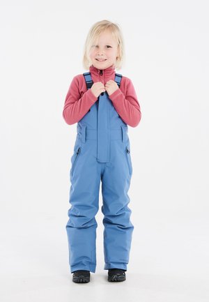Blauwe geïsoleerde sneeuwoverall met verstelbare schouderbanden, zijzakken met ritssluiting en elastische manchetten. Draag het over een rood fleece shirt. Zwarte laarzen.