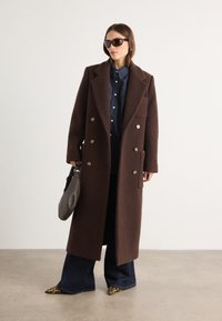 Cappotto di lana marrone, design doppiopetto, grandi rever, abbinato a una camicia di jeans, jeans a zampa d'elefante e scarpe con stampa leopardata. Tiene una borsa grigia.