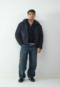Schwarze Daunenjacke mit Kapuze, kombiniert mit einem dunklen Pullover und einem hellrosa Hemd, ergänzt durch locker sitzende blaue Jeans und schwarze Sneakers.