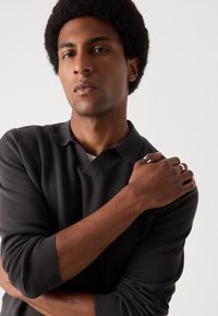 Crna polo majica s teksturiranim pletenjem, s v-izrezom. Model ima afro frizuru i nosi srebrne prstenove na više prstiju.