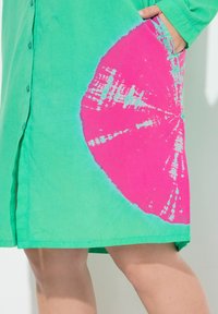 Robe chemise en lin vert avec un grand motif de tie-dye circulaire rose. Boutonnage à l'avant et poches sur les côtés. Texture douce.