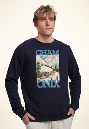 Man draagt een marineblauwe trui met een afbeelding van een besneeuwde berghut en skiërs, met de tekst 'CHAMONIX', handen in de zakken, kijkt iets naar rechts.