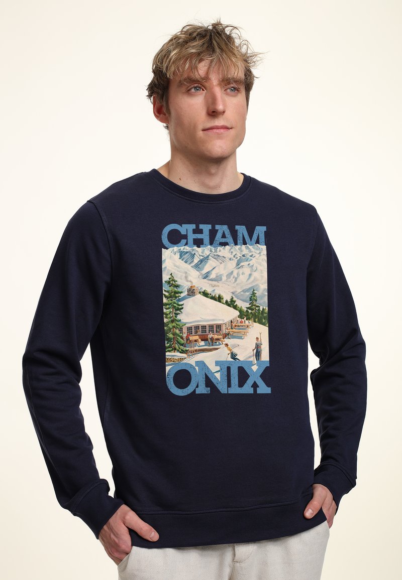 Hombre con sudadera azul marino con gráfico de una cabaña en la montaña nevada y esquiadores, con texto "CHAMONIX", manos en los bolsillos, mirando ligeramente a la derecha.