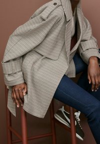 Geruite oversized jas in beige en zwarte stof, met een gestructureerde kraag, opgerolde mouwen en een losse pasvorm over donkere denim.