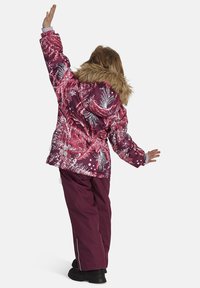 Winterjack van een kind in een paarse, gestreepte stof met een nep bont capuchon, gecombineerd met donkere paarse broeken en zwarte laarzen, van achteren gezien.