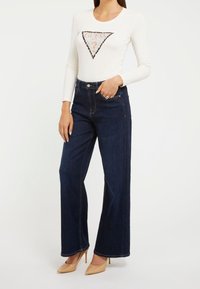 Top bianco a maniche lunghe con logo a triangolo in paillettes, abbinato a jeans a vita alta bleu scuro e scarpe con tacco nudo.