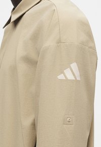Μανίκι μπεζ σακακιού με λευκό λογότυπο Adidas και μικρή τετράγωνη λεπτομέρεια κουμπιού κοντά στον αγκώνα, που δείχνει υφασμένο ύφασμα.