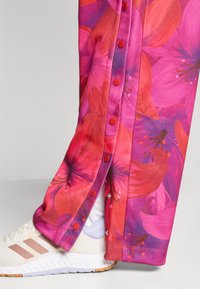 Πολύχρωμο floral παντελόνι από ζωηρό ροζ ύφασμα με μεγάλα κόκκινα και μωβ λουλούδια, διακοσμημένο με κόκκινα κουμπι ά κατά μήκος της πλαϊνής ραφής. Φορεμένο με λευκά αθλητικά παπούτσια.