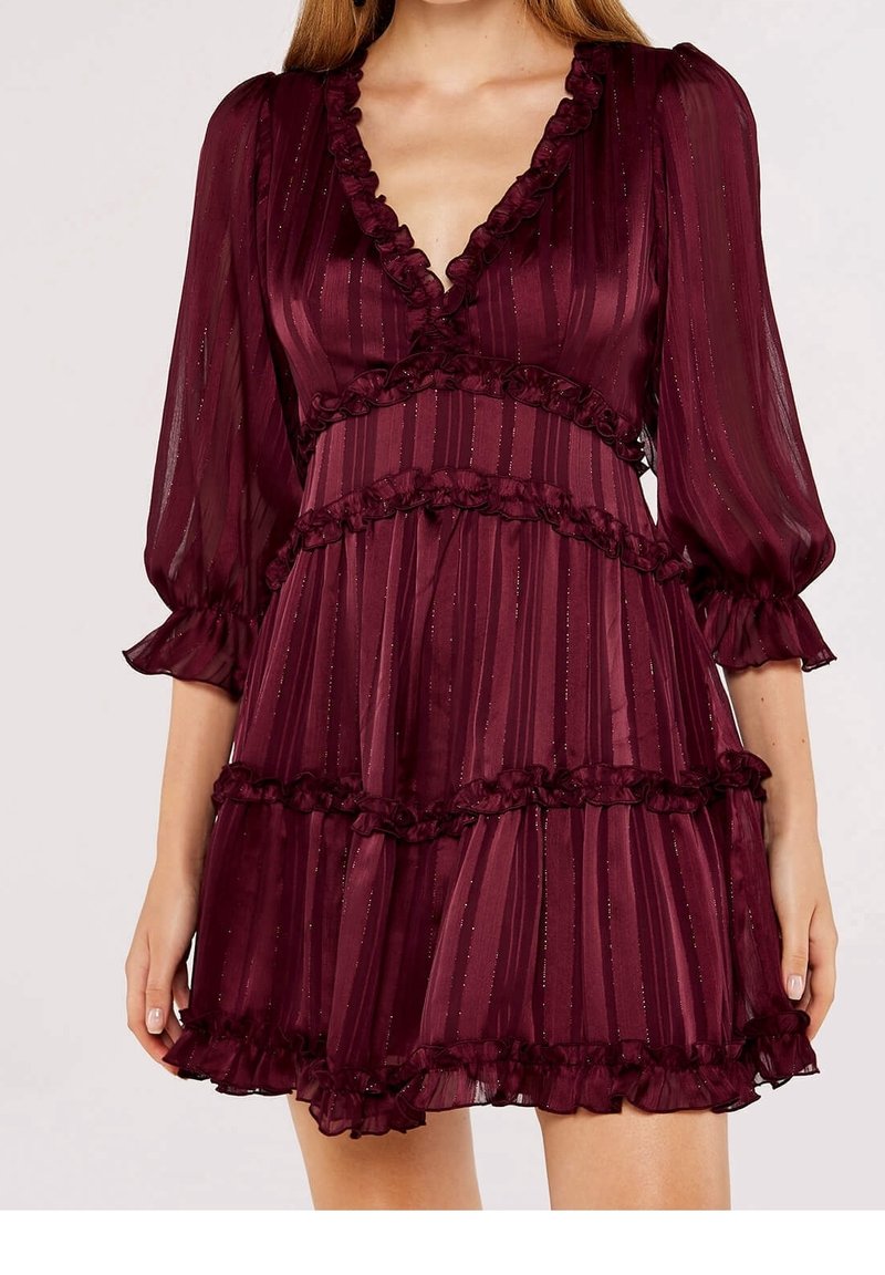 Femme portant une robe bordeaux à manches longues, col en V, avec des volants et de fines rayures verticales, terminant au-dessus des genoux.