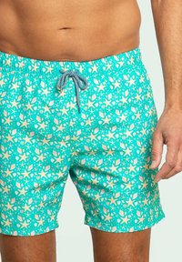 Bañador turquesa con un estampado de estrellas de mar y conchas, cintura elástica con cordón azul y blanco, textura de tela suave.