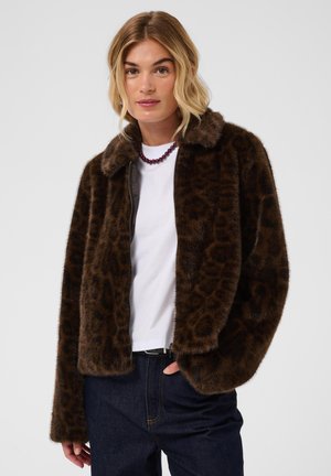 Femme aux cheveux blonds portant une veste en fausse fourrure imprimé léopard marron foncé, une chemise blanche, un jean foncé et un collier de perles violet, debout sur un fond blanc.
